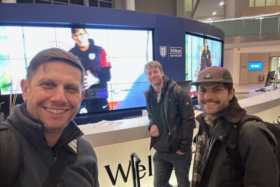 Three crew members selfie in front of a large video display wall