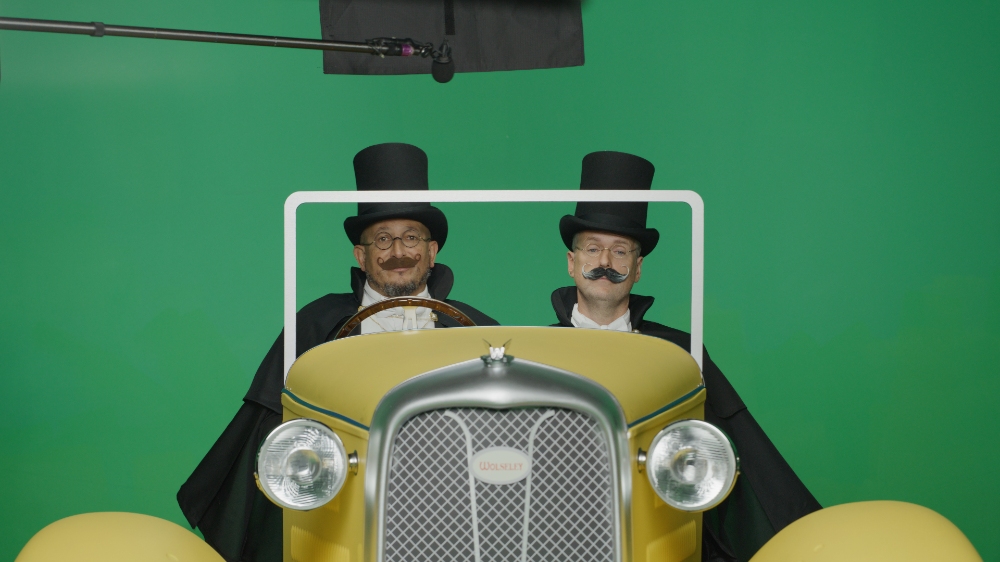 Two men in top hats and mustaches in vintage yellow car on green screen