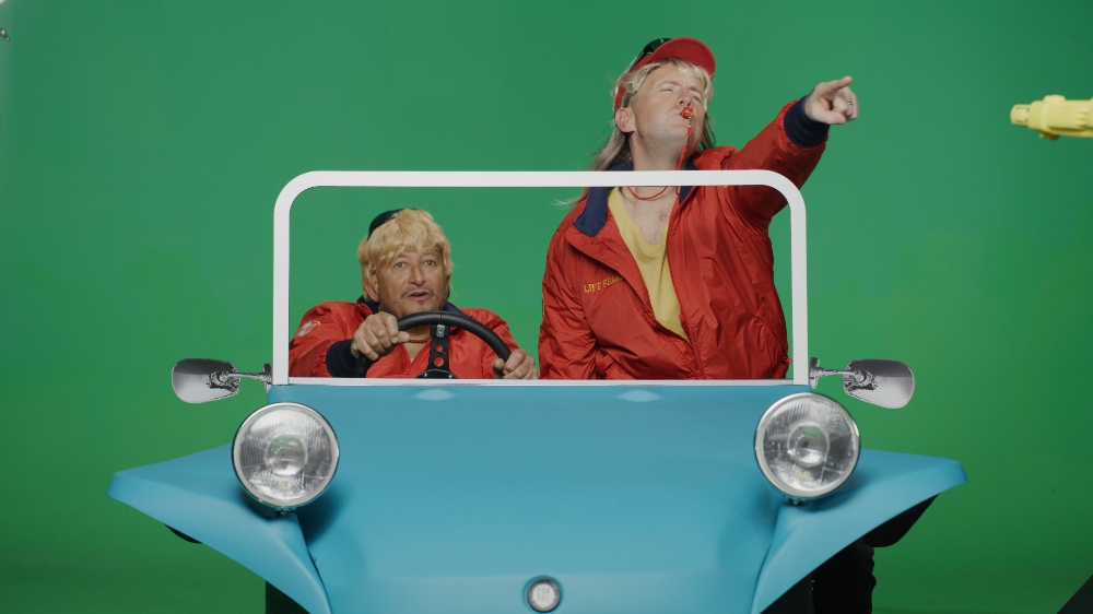 Two presenters in blue prop car pointing on Car SOS green screen