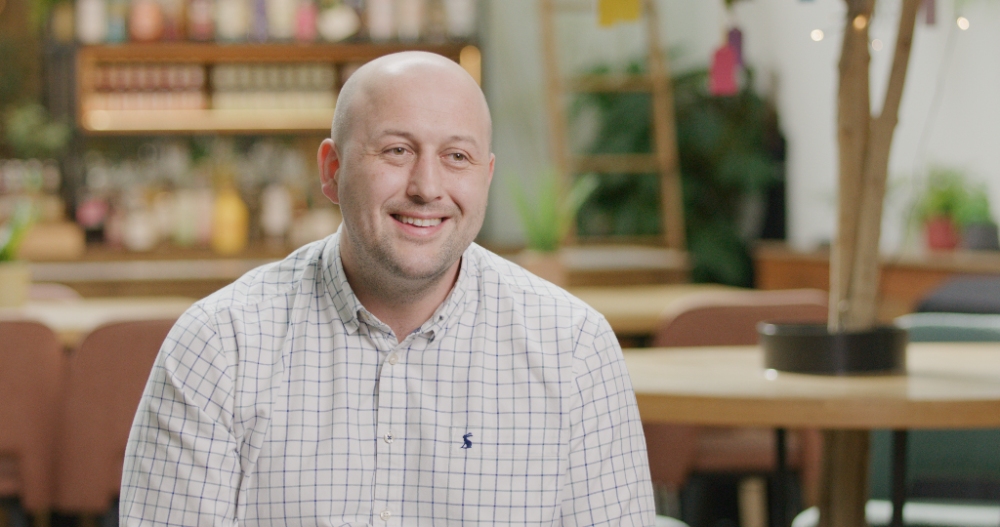 Smiling man in white shirt speaking on camera in café setting