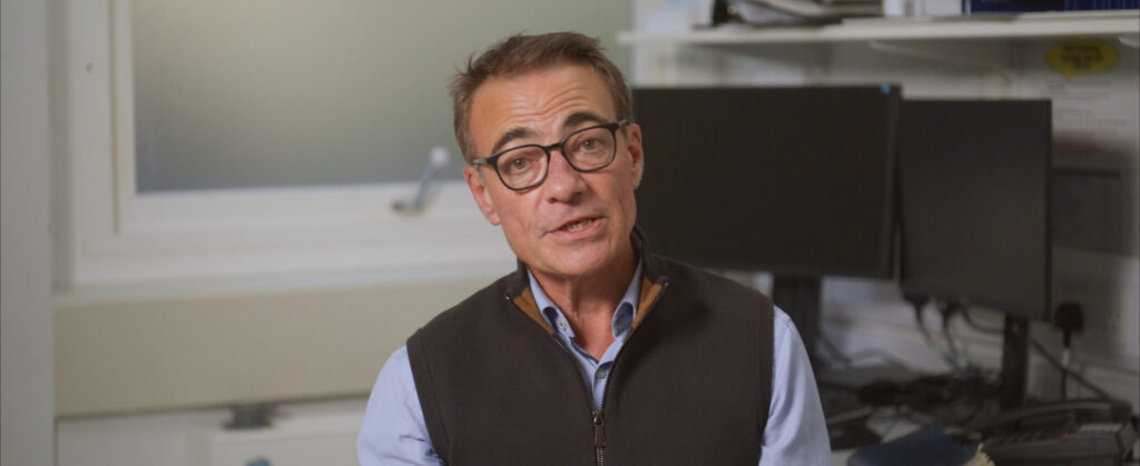 Man in vest and glasses speaking on camera in an office